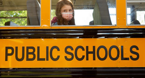 A single student, wearing a face mask, is sitting on a school bus and looking through the window.