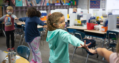 Young students wearing face masks stand in a line with their arms up to ensure they are socially distanced.