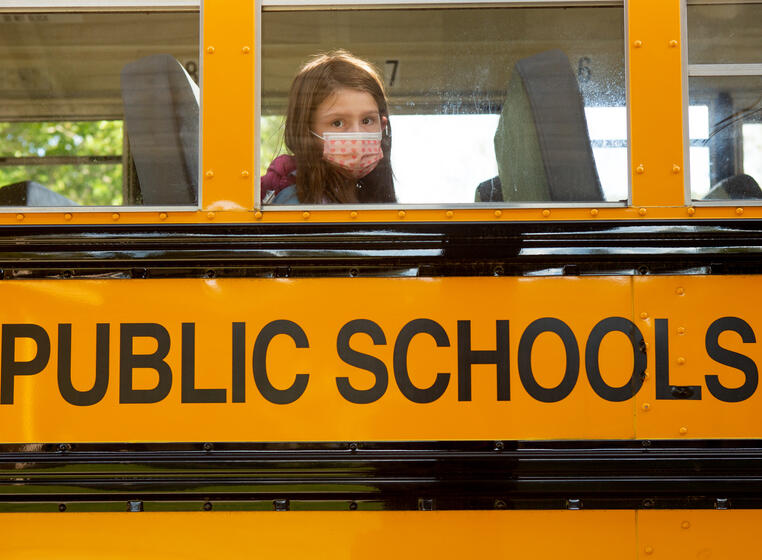 A single student, wearing a face mask, is sitting on a school bus and looking through the window.