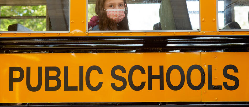 A single student, wearing a face mask, is sitting on a school bus and looking through the window.