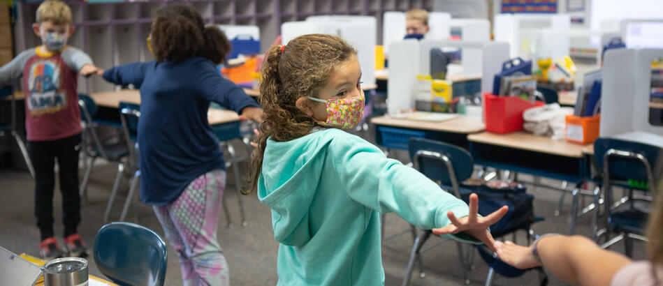 Young students wearing face masks stand in a line with their arms up to ensure they are socially distanced.