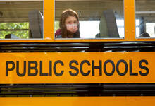 A single student, wearing a face mask, is sitting on a school bus and looking through the window.