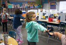 Young students wearing face masks stand in a line with their arms up to ensure they are socially distanced.